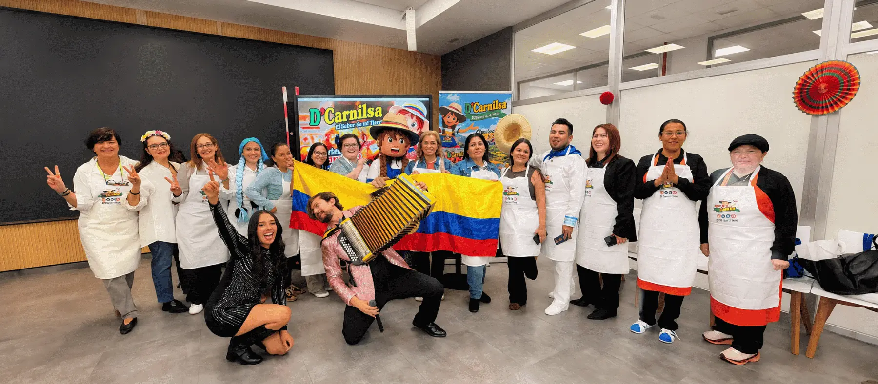 Eventos D'Carnilsa 5 Participantes con delantales de D'Carnilsa, la mascota de la marca, un acordeonista y la bandera de Colombia en la Escuela Municipal de Hostelería y Alimentación de Madrid